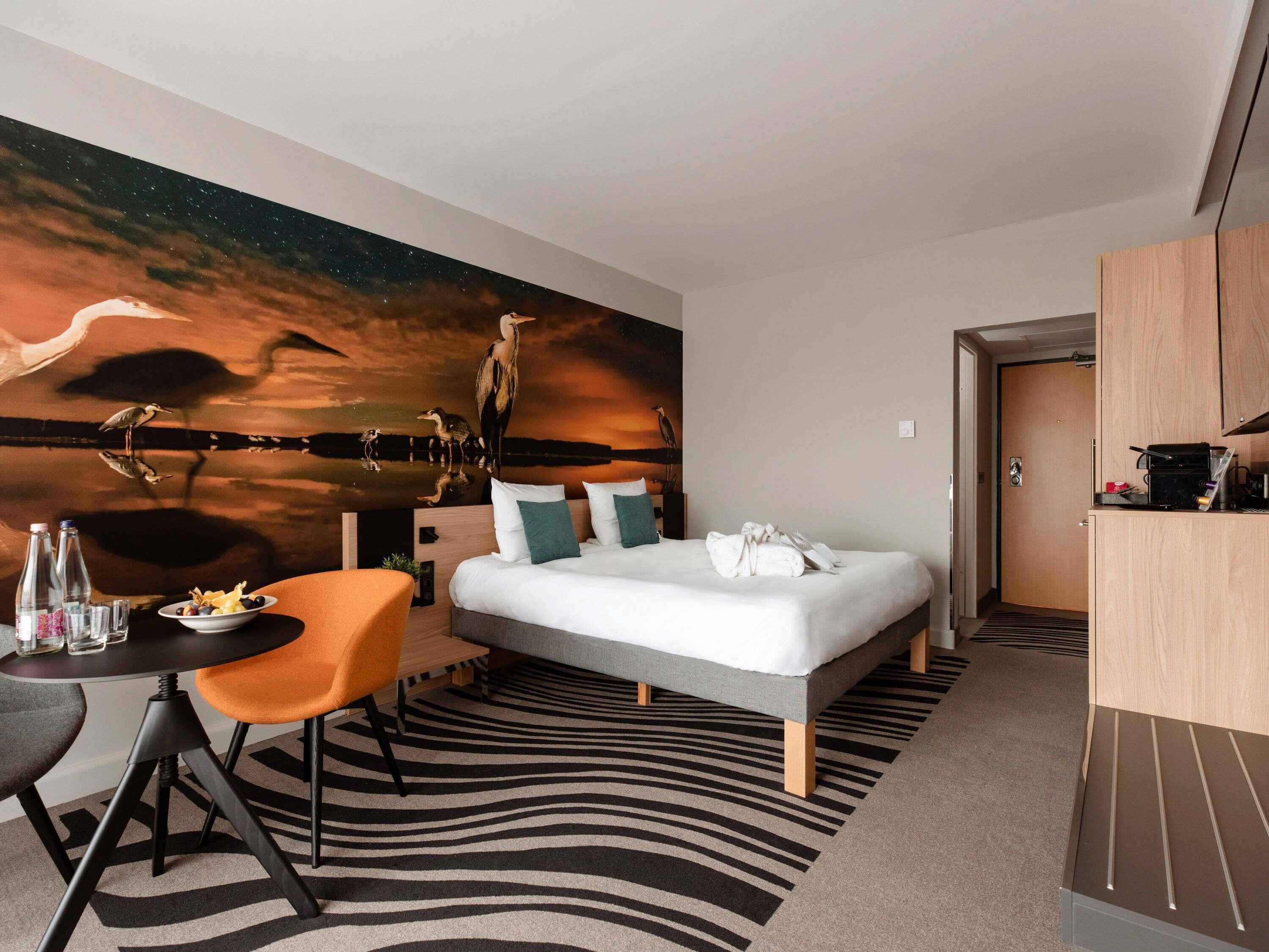 Una habitación de hotel con una cama, un escritorio con silla, un mural en la pared y un armario.