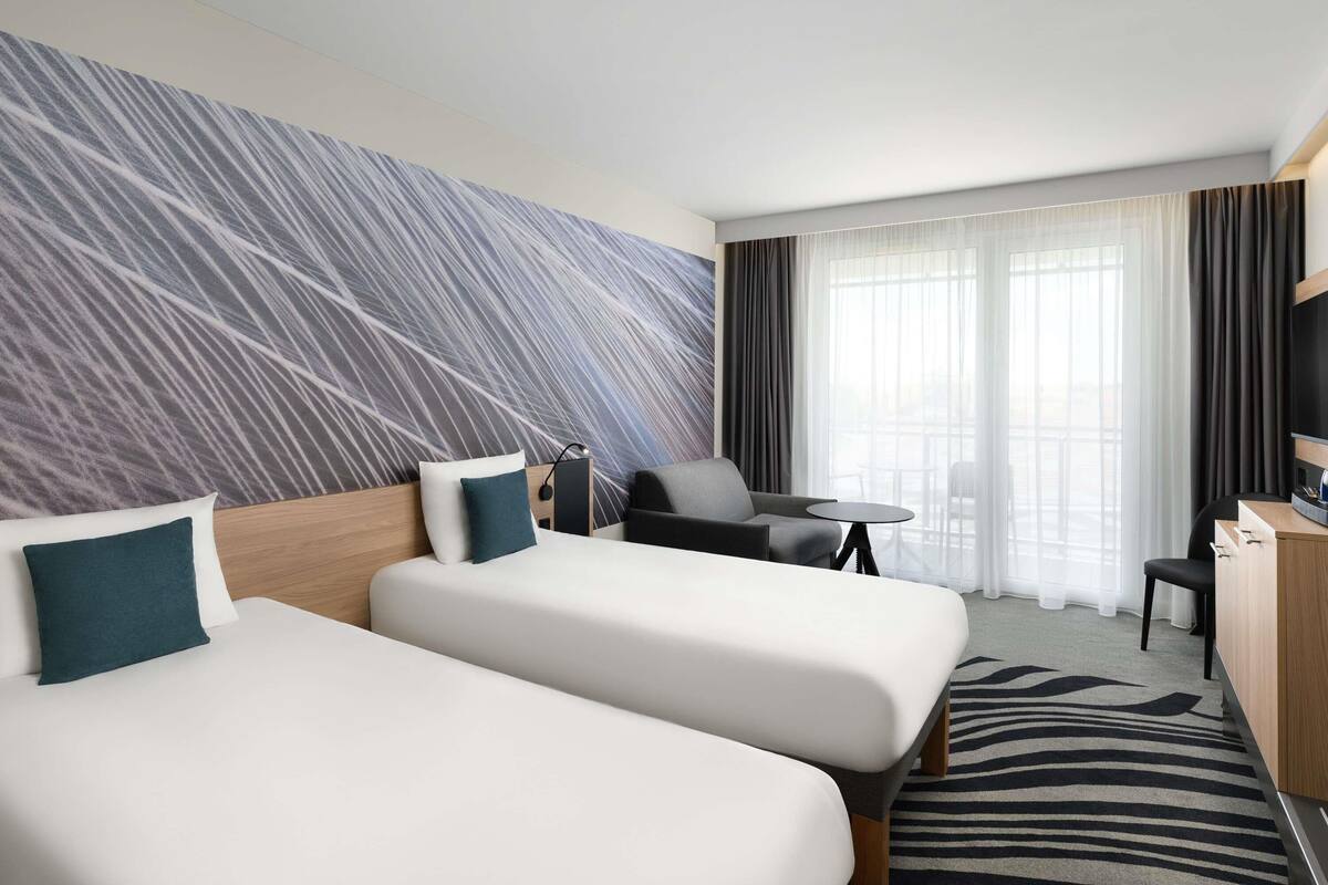 A hotel room with two beds, a desk, a chair, a TV, and a large window with curtains.