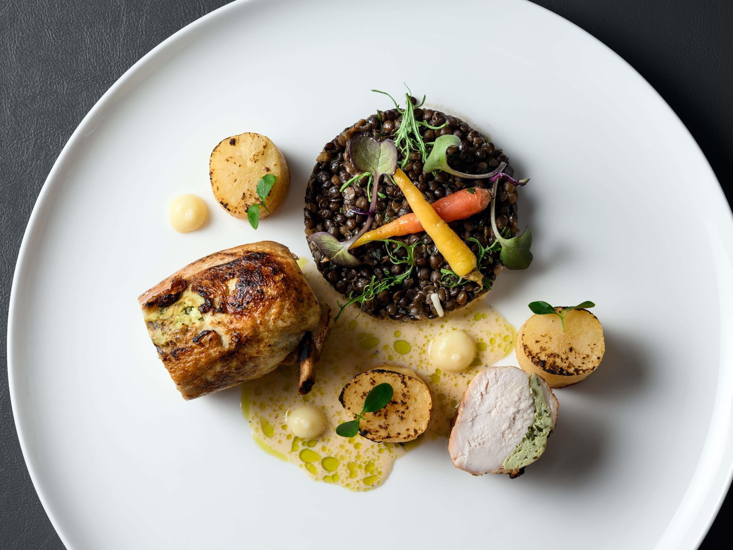 A white plate with a variety of food items including a grilled chicken breast, a mound of black lentils, and several small round vegetables.