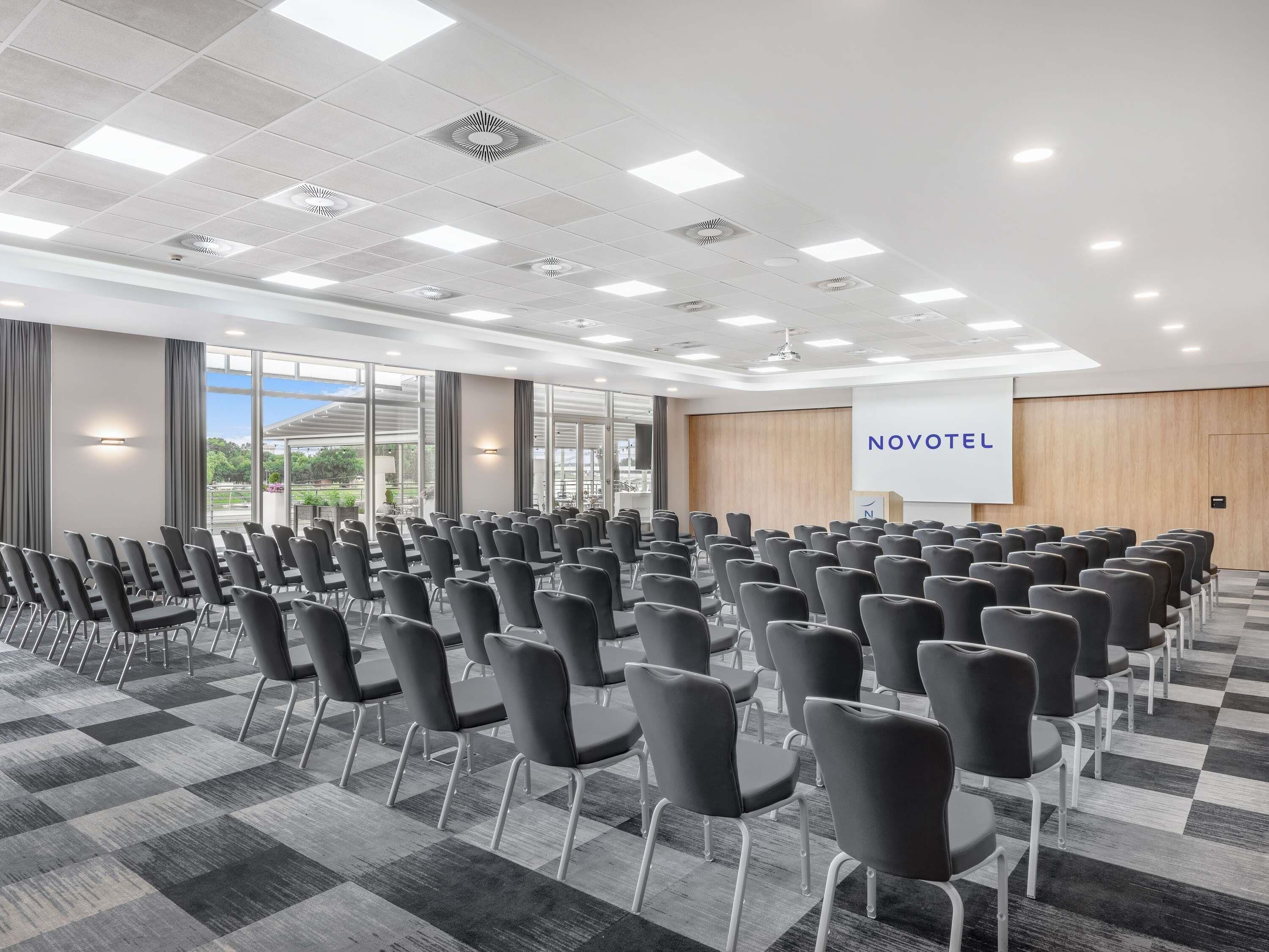 A conference room with rows of chairs, a projector screen, and a 'NOVOTEL' sign.