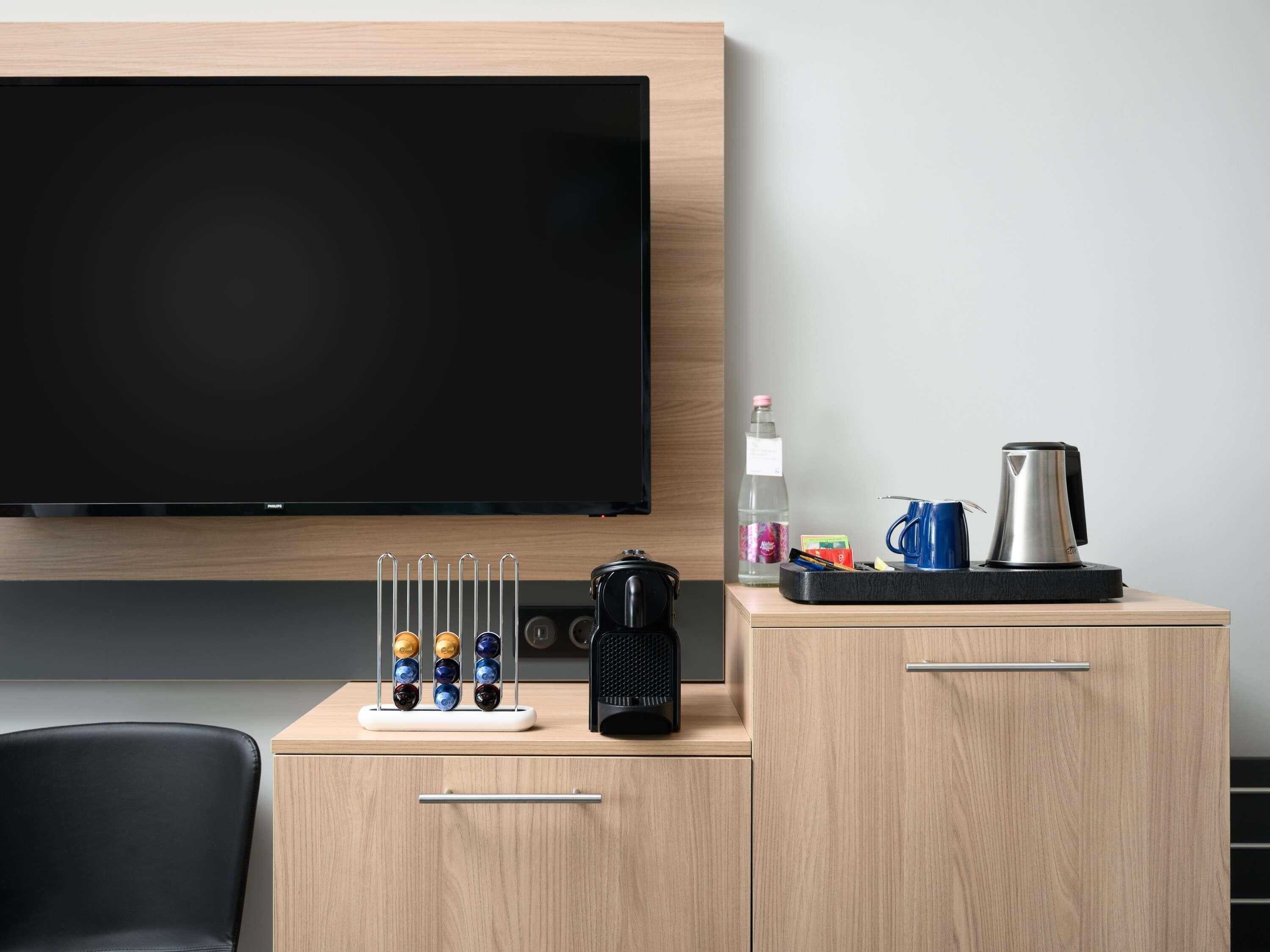 A hotel room with a flat-screen TV mounted on the wall, a coffee maker, a water bottle, and a set of colorful cups on a wooden cabinet.