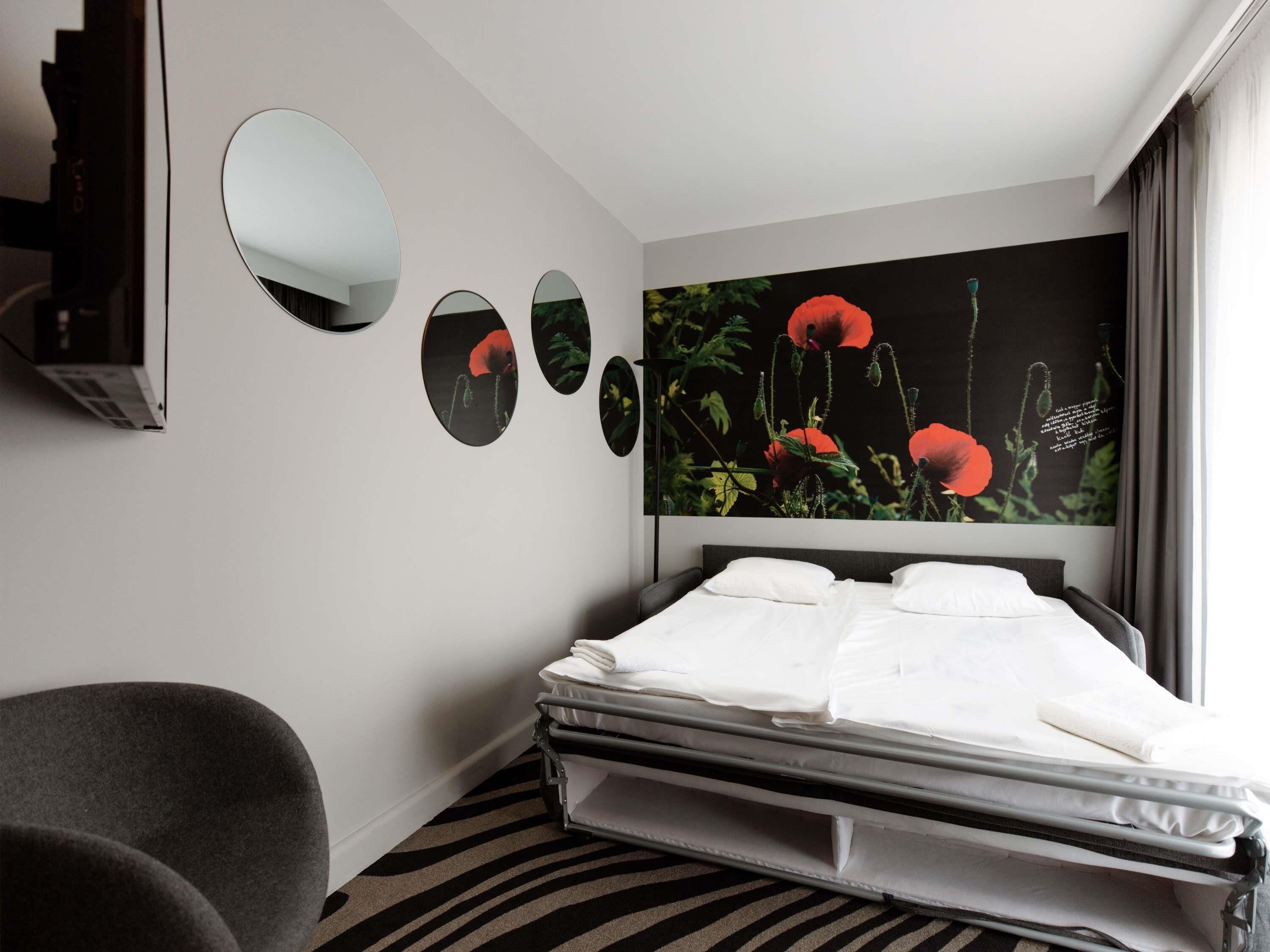 Ett modernt hotellrum med en säng, en stol, en spegel som hänger på väggen och ett stort konstverk med blommotiv.