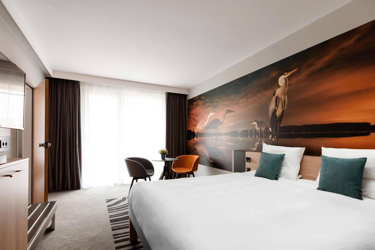 A hotel room with a large bed, a desk, a chair, a wardrobe, and a wall mural.