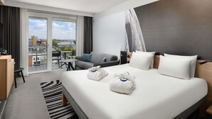 A modern hotel room with a large bed, a desk, a chair, and a balcony with a view of a river and buildings. - Novotel Szeged (Szeged)