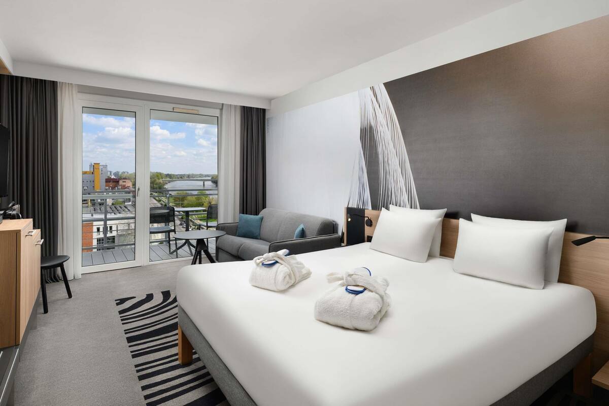 A modern hotel room with a large bed, a desk, a chair, and a balcony with a view of a river and buildings.