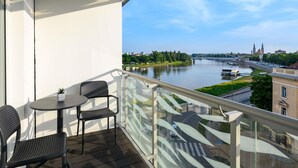 A balcony with a table and chairs, overlooking a river with a bridge and buildings in the background. - Novotel Szeged (Szeged)