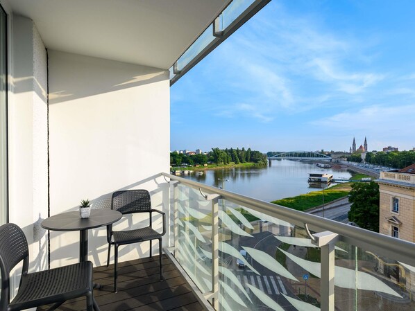 A balcony with a table and chairs, overlooking a river with a bridge and buildings in the background. - Novotel Szeged (Szeged)