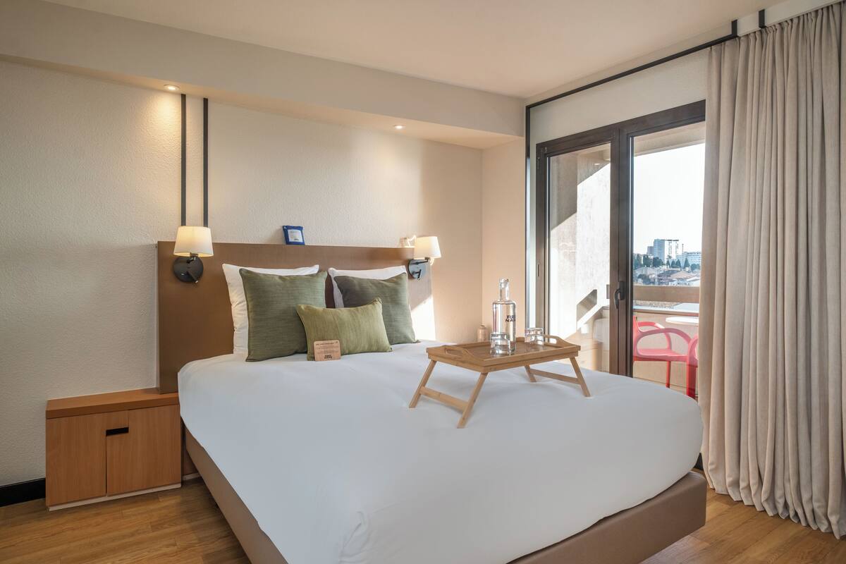 A hotel room with a large bed, a bedside table, a nightstand, a window with curtains, and a view of the city.