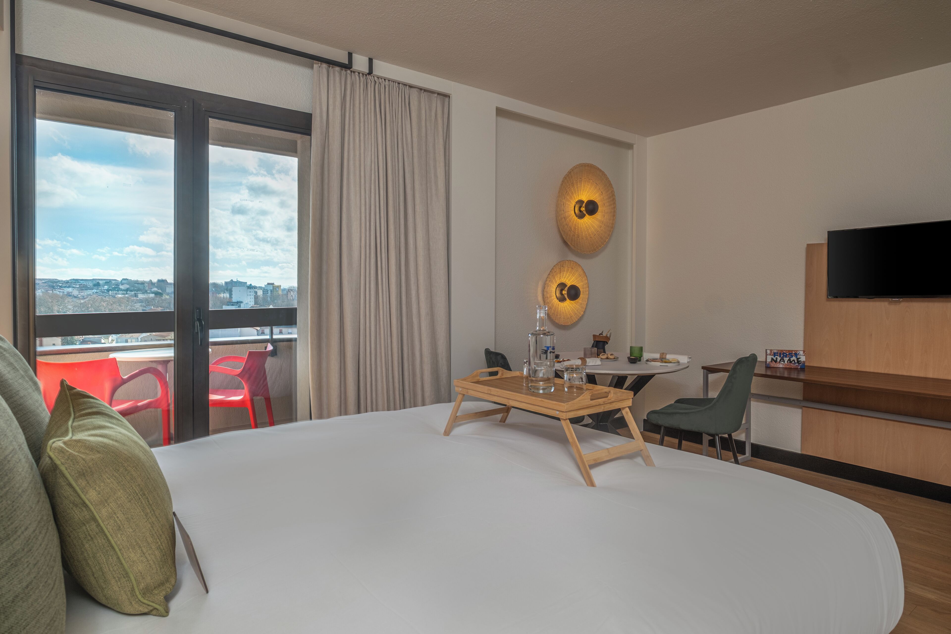 A hotel room with a large bed, a dining table with chairs, a TV, and a view of the city through large windows.
