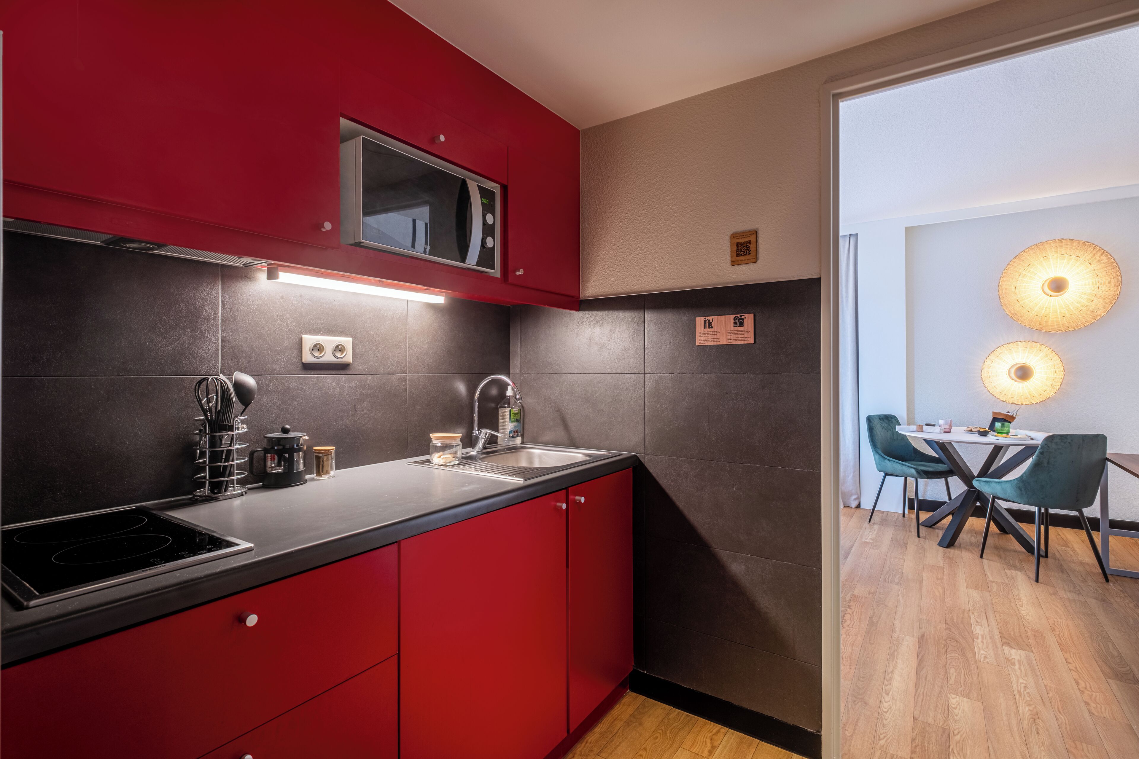 A modern kitchen with red cabinets, a black countertop, and a built-in microwave. There is a dining area with a table and chairs visible through an open doorway.