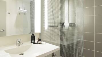 Un baño moderno con un espejo grande, un lavabo blanco, cabina de ducha de vidrio y un cubo de metal.