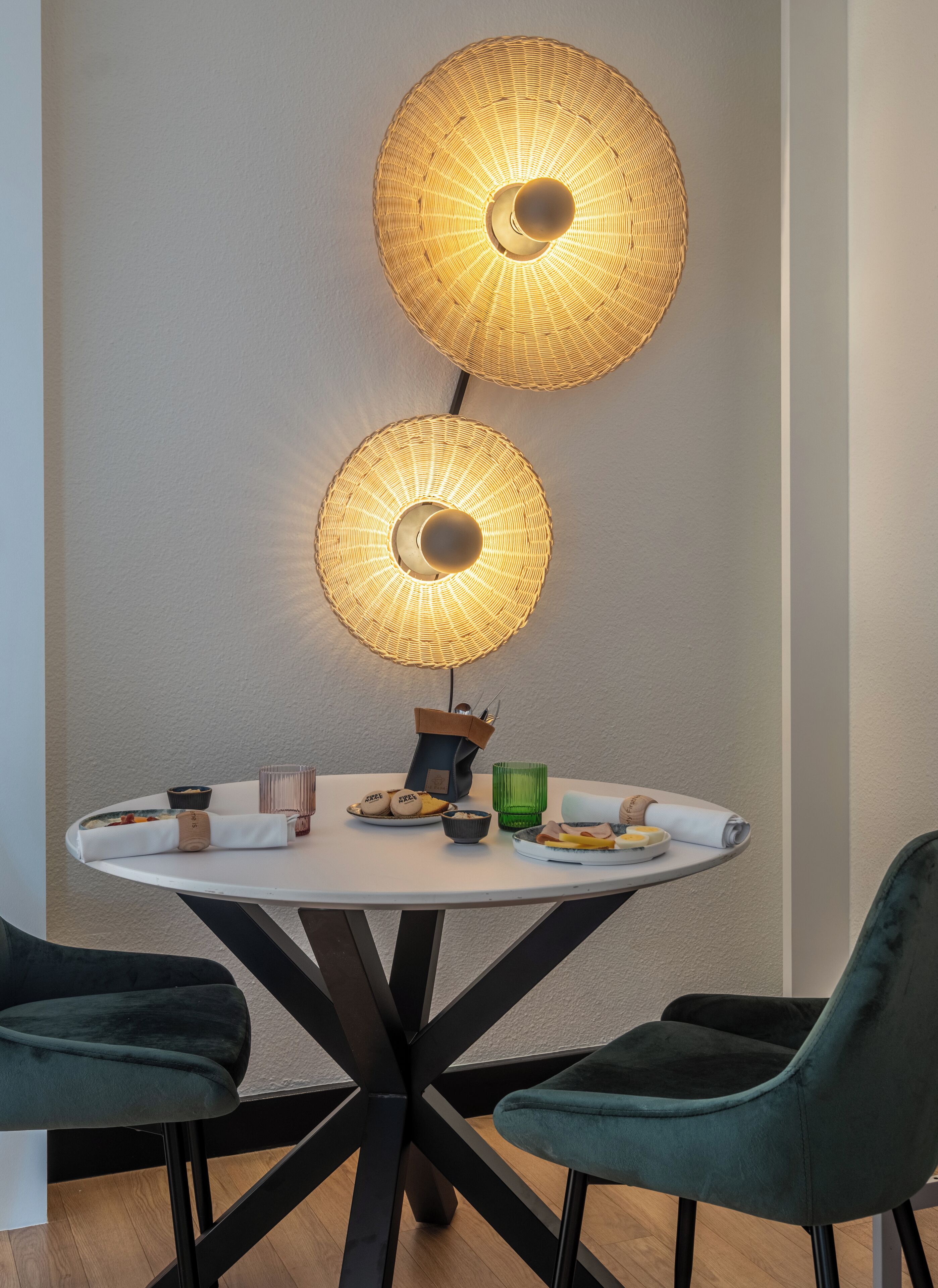 Ett runt matbord med en vit bordsskiva och svarta metallben, omgivet av två mörkgröna fåtöljer klädda i sammet. Ovanför bordet hänger två stora, runda lampor av flätat material, med en central ljuskälla.