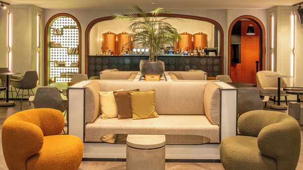 Eine moderne Lobby mit hoher Decke, einer zentralen Sitzecke mit Sofas und Sesseln, einer großen Pflanze und einer Bar mit Regalen voller Flaschen.
