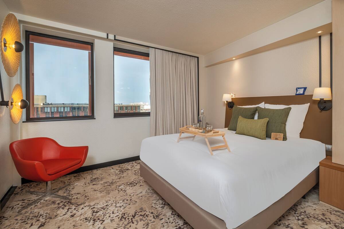 A hotel room with a large bed, a red chair, a window with curtains, and a tray with amenities.