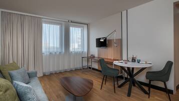 Una sala de estar moderna con un sofá, una mesa de centro, una mesa de comedor con sillas, un televisor montado en la pared y ventanas grandes con cortinas.