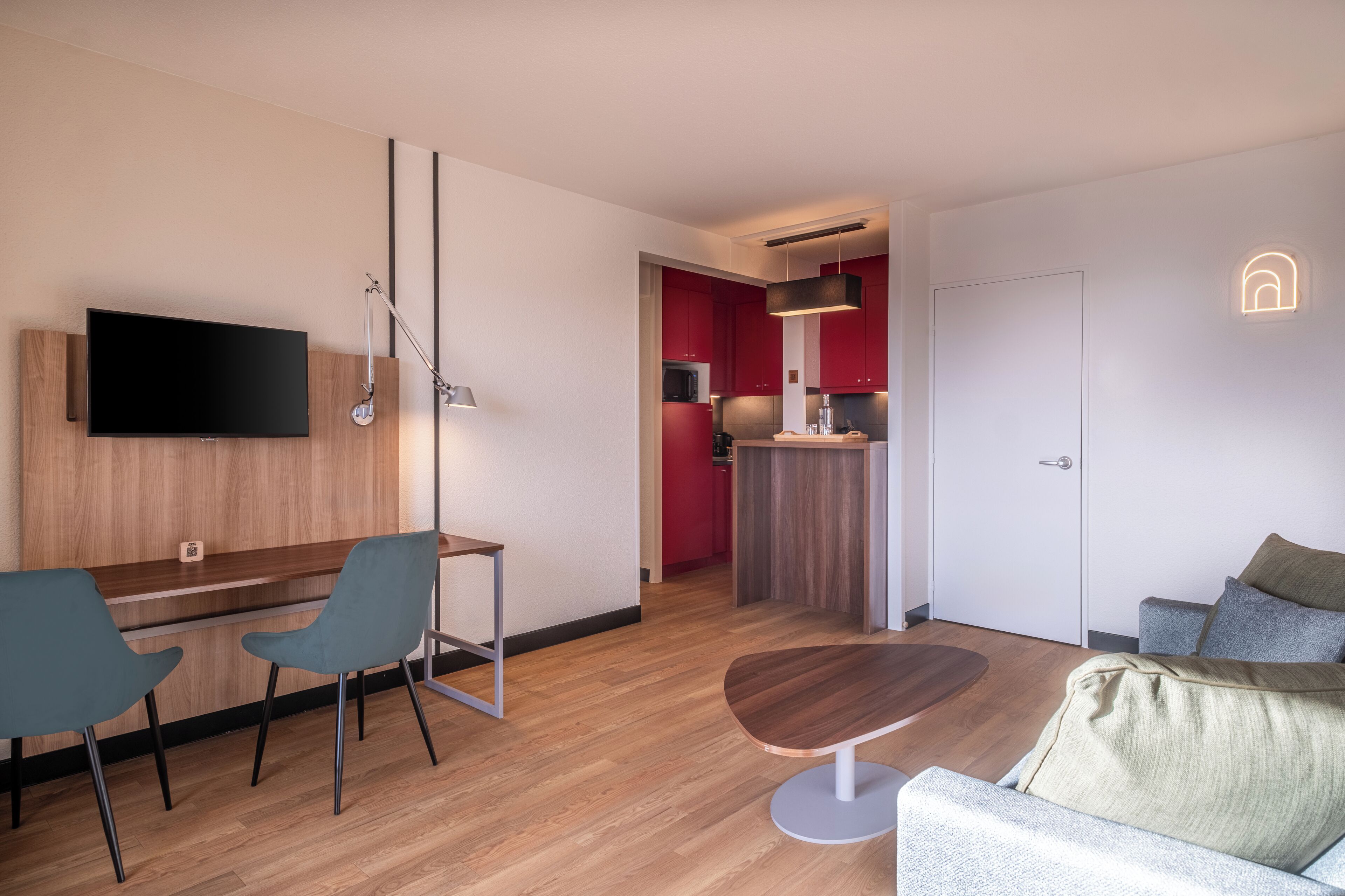 A modern hotel room with a wooden floor, a flat-screen TV mounted on the wall, a dining area with two chairs, a sofa, and a small table.