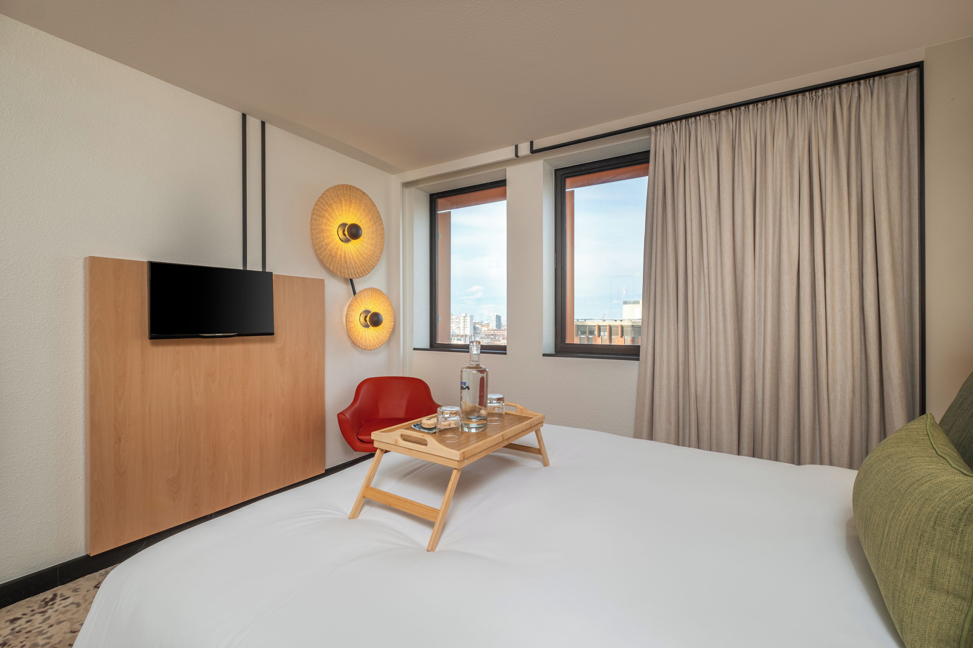 A hotel room with a bed, a TV, a wooden side table with a tray, a red chair, and a city view through the windows.