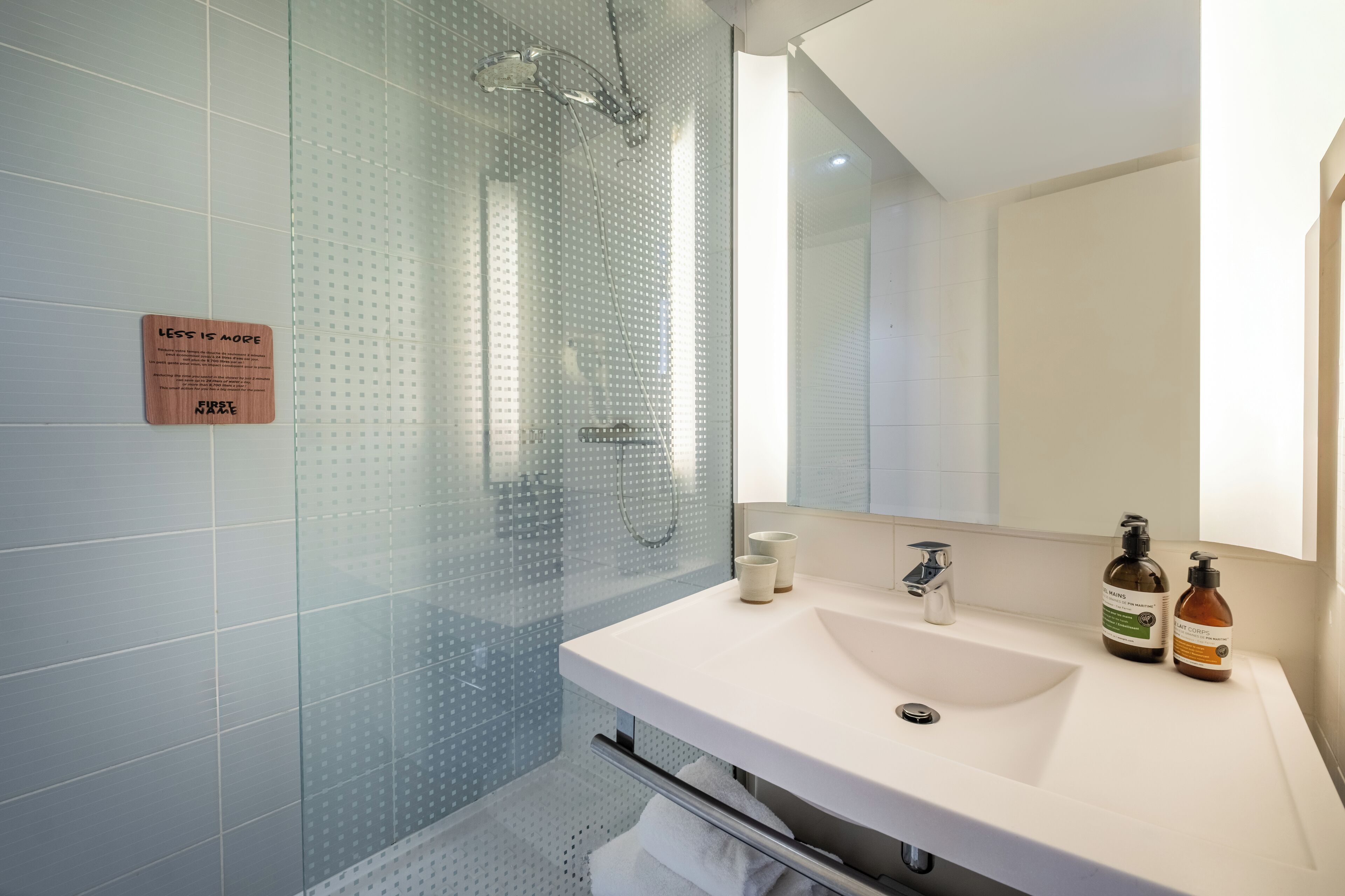 A modern bathroom with a glass shower enclosure, a white sink with two bottles of shampoo, and a sign on the wall.