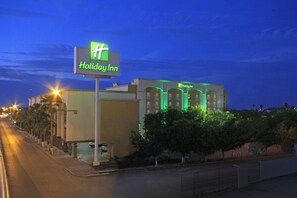 A Holiday Inn hotel with a prominent green sign, a row of palm trees along the street, and a well-lit facade. - Holiday Inn Monclova by IHG (Monclova)