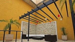 Un patio techado con pared de piedra, un banco blanco acolchado y una estructura de pérgola negra.