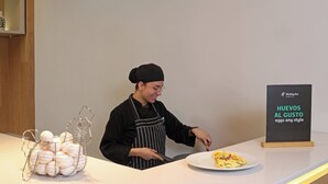 Una persona con delantal detrás de una barra, con un plato de comida y un letrero que dice 'Holiday Inn HUEVOS AL GUSTO eggs any style'.