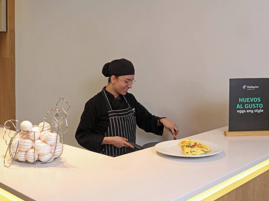 A person in an apron at a counter with a plate of food and a sign that reads 'Holiday Inn HUEVOS AL GUSTO eggs any style.'
