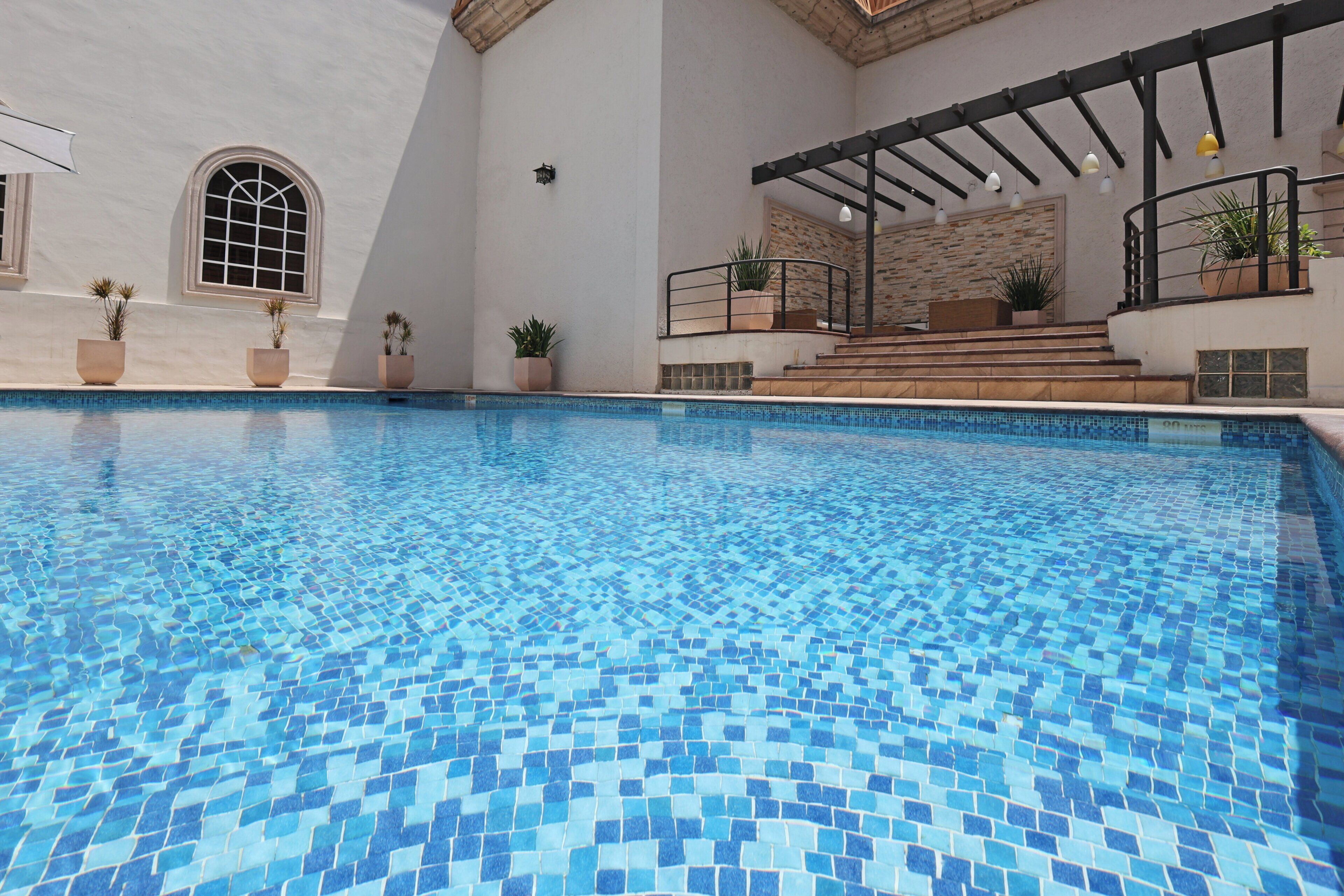 A swimming pool with a tiled floor, surrounded by potted plants and a covered area with steps leading up to a balcony.