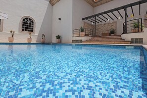 Piscina com piso ladrilhado, rodeada por plantas em vasos e uma área coberta com escadas que levam a uma varanda.