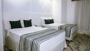 Habitación de hotel con dos camas, cada una con ropa de cama blanca y mantas a rayas. Se aprecia un cabecero de madera, una pared blanca y una ventana con cortinas blancas.