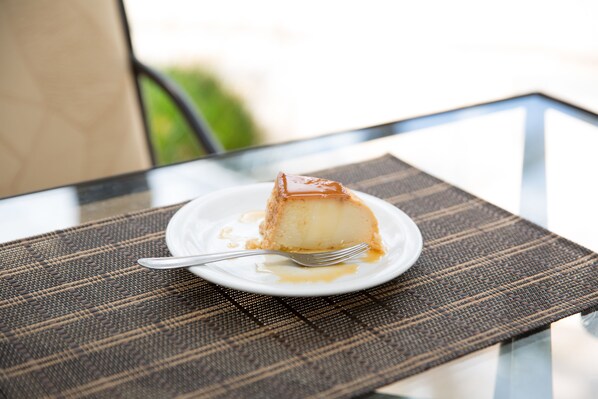 Una porción de flan en un plato blanco, con un tenedor sobre una mesa de vidrio con un mantel tejido.