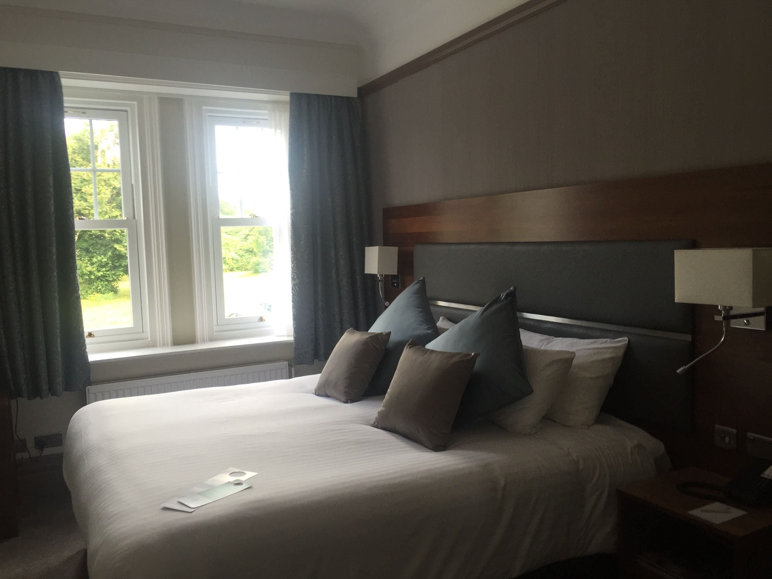 Una habitación de hotel con una cama grande, una ventana con cortinas y una mesita de noche con lámpara.