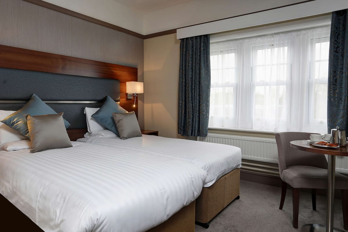 Une chambre d’hôtel avec un grand lit, une table de chevet avec une lampe, une petite table avec une tasse et une soucoupe, et une fenêtre avec des rideaux.