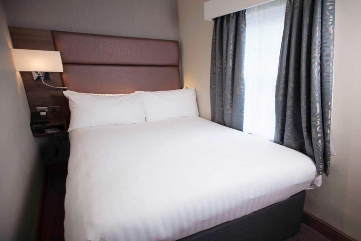 Une chambre d’hôtel avec un grand lit, une table de chevet, une lampe et une fenêtre avec des rideaux.