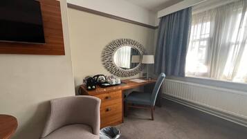 A hotel room with a wooden desk, a chair, a television, a mirror, a lamp, and a window with curtains.