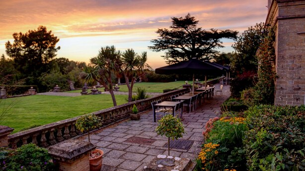 An outdoor patio with stone flooring, a row of tables and chairs, and a view of the sky at sunset.