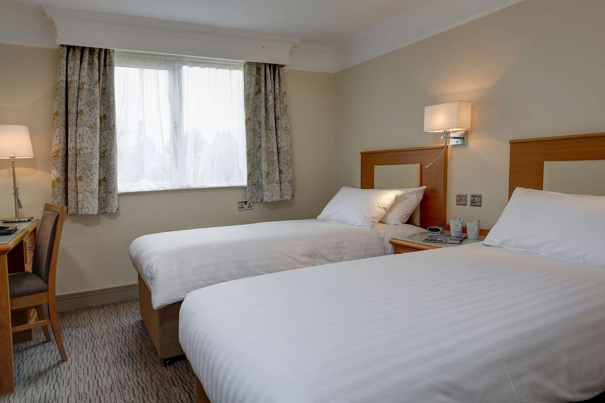 Une chambre d’hôtel avec deux lits simples, un bureau avec une chaise, une lampe et une fenêtre avec des rideaux.
