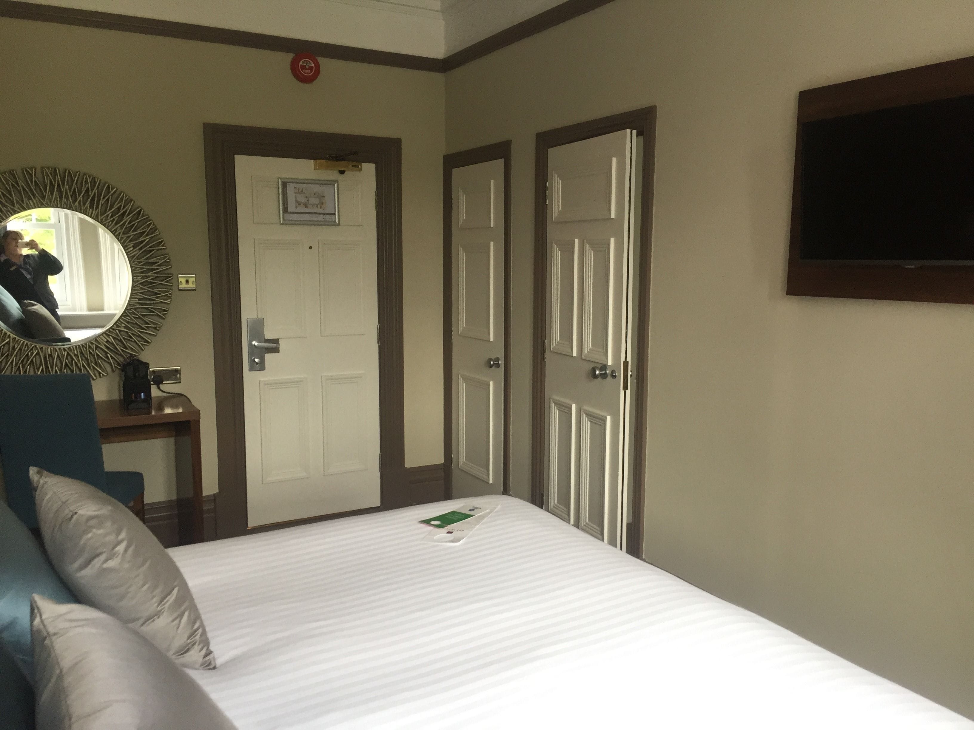 A hotel room with a bed, a desk with a coffee maker, a mirror, and two doors.