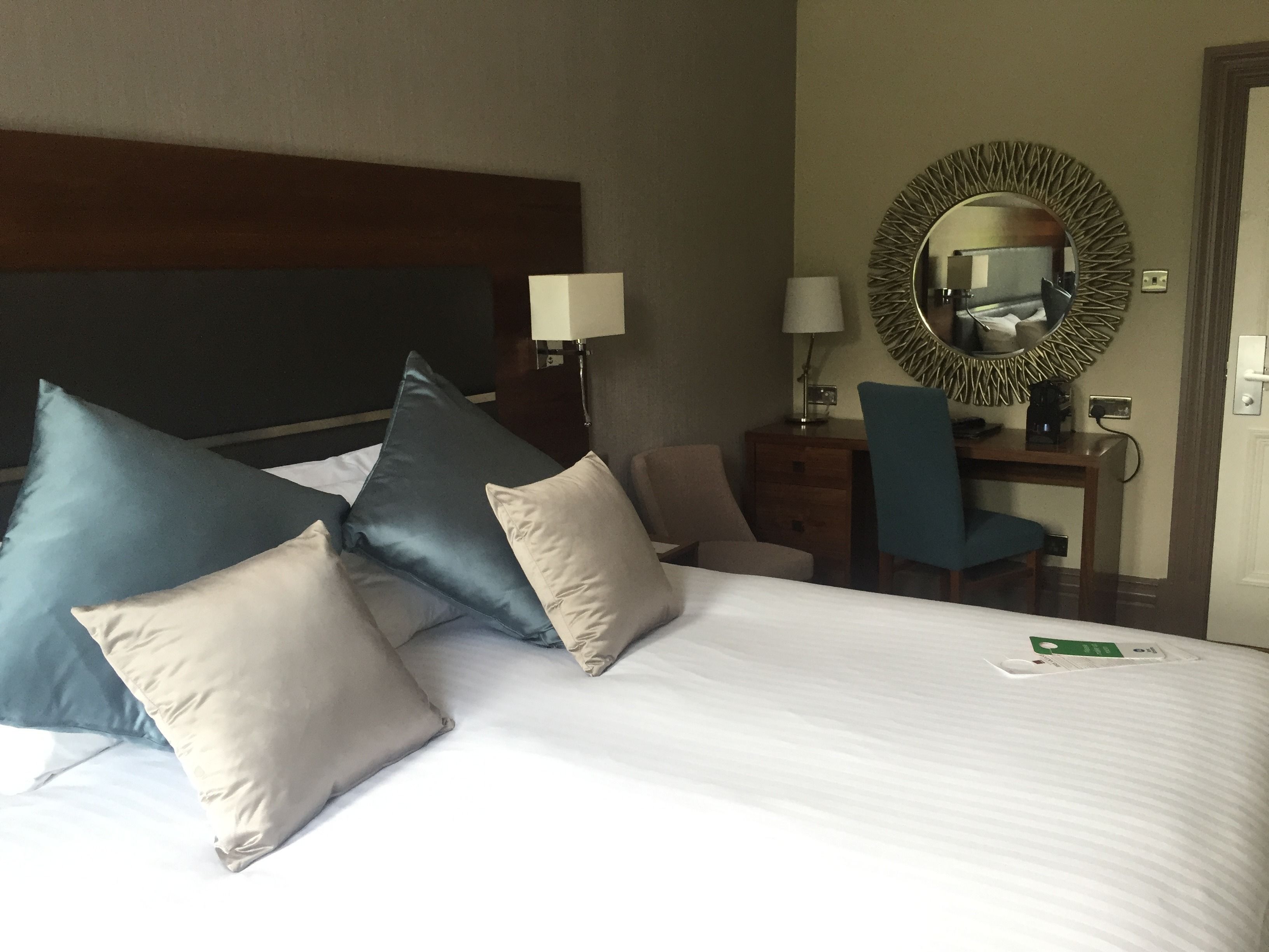 Une chambre d’hôtel avec un grand lit, plusieurs oreillers, un bureau avec une chaise, un miroir et une lampe.