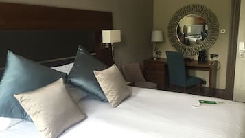 A hotel room with a large bed, multiple pillows, a desk with a chair, a mirror, and a lamp.