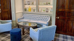 A room with a tufted blue sofa, two matching armchairs, a glass coffee table, and a patterned rug. A wooden door and a shelf with decorative items are visible.