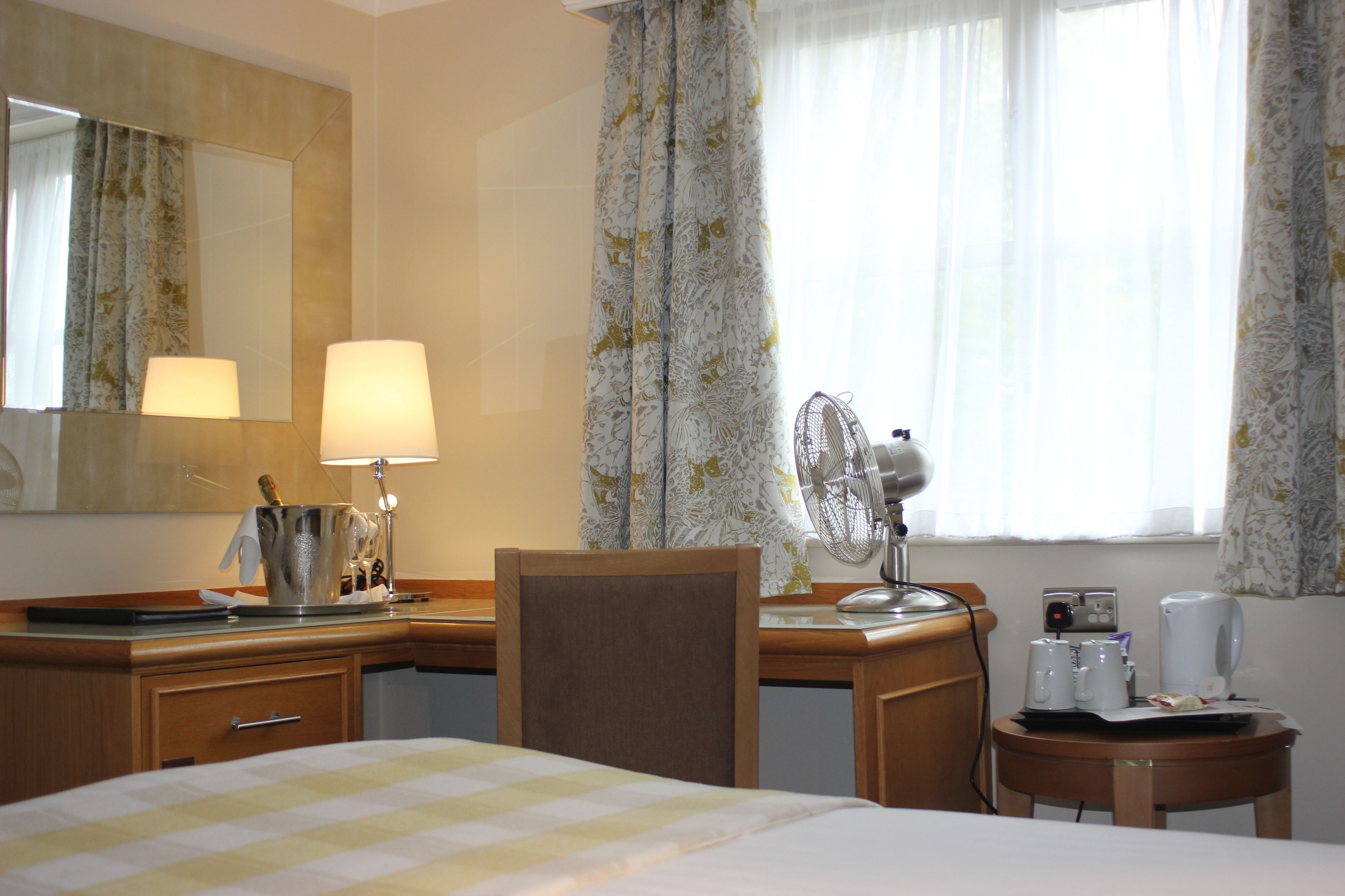 A hotel room with a bed, a desk with a lamp, a fan, a coffee maker, and a mirror.