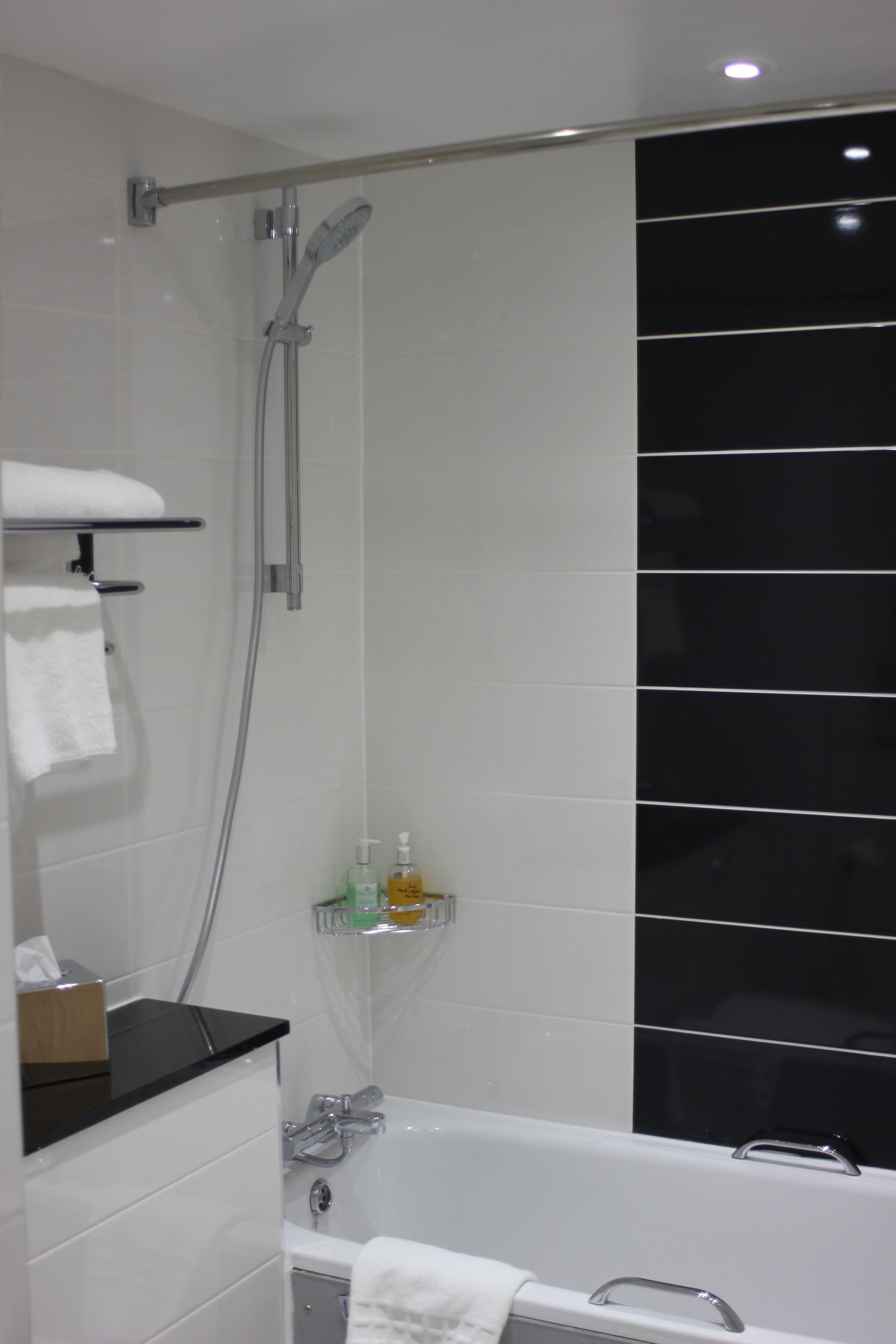 A modern bathroom with a shower, a bathtub, and a toilet. There are white tiles on the walls and a black tiled accent wall. A towel is hanging on a rack, and there are toiletries on a shelf.