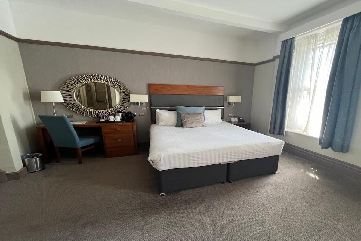 Une chambre d’hôtel avec un lit, un bureau, une chaise et une grande fenêtre avec des rideaux.