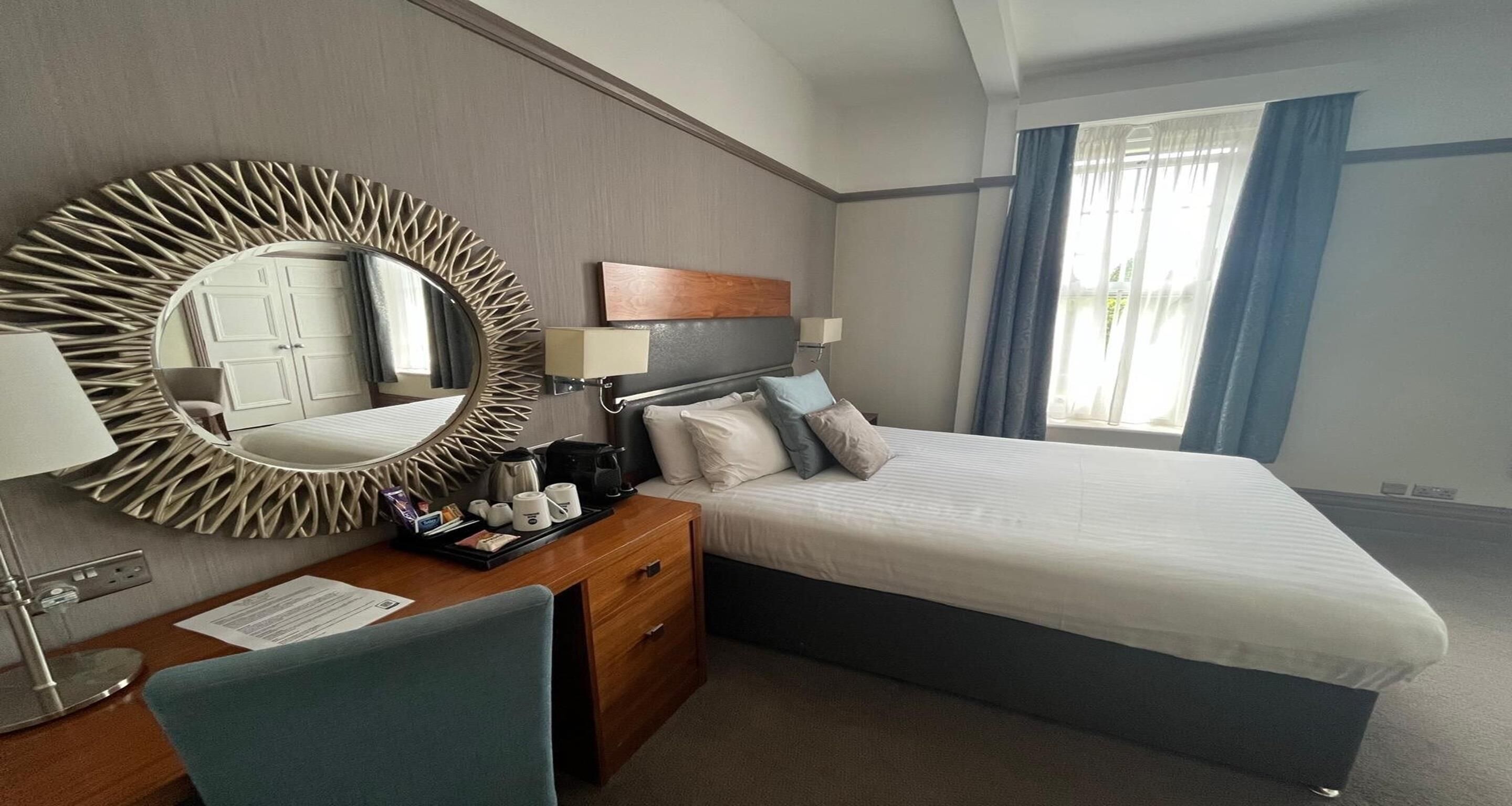 Une chambre d’hôtel comprenant un lit, un bureau avec une chaise, un grand miroir, une tête de lit en bois et une fenêtre avec des rideaux.