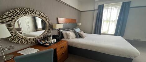 Une chambre d’hôtel comprenant un lit, un bureau avec une chaise, un grand miroir, une tête de lit en bois et une fenêtre avec des rideaux.
