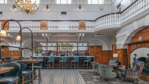 A spacious bar area with high ceilings and a chandelier, featuring a bar counter with stools and a seating area with chairs.