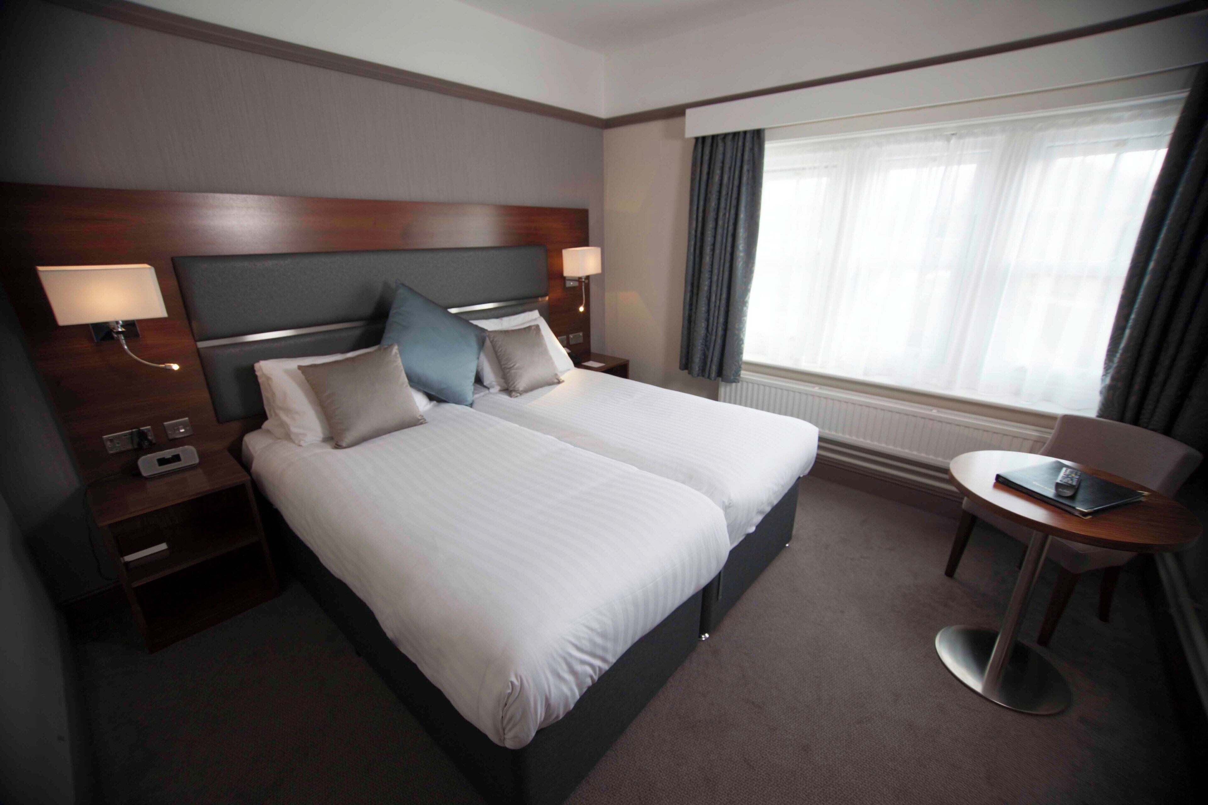 A hotel room with a large bed, two bedside tables with lamps, a small round table, and a window with curtains.