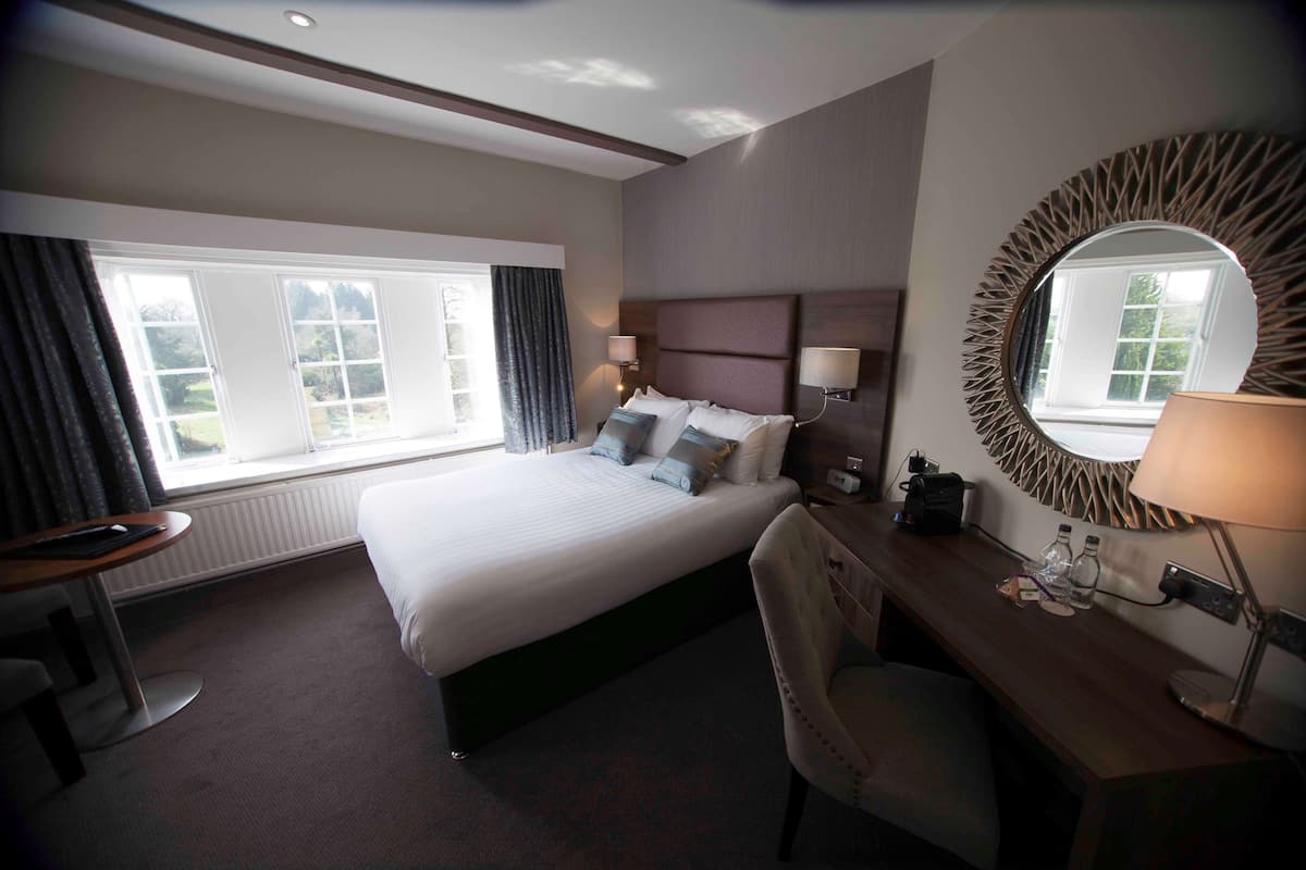 Une chambre d’hôtel avec un grand lit, un bureau avec une chaise, un miroir rond et une fenêtre avec des rideaux.