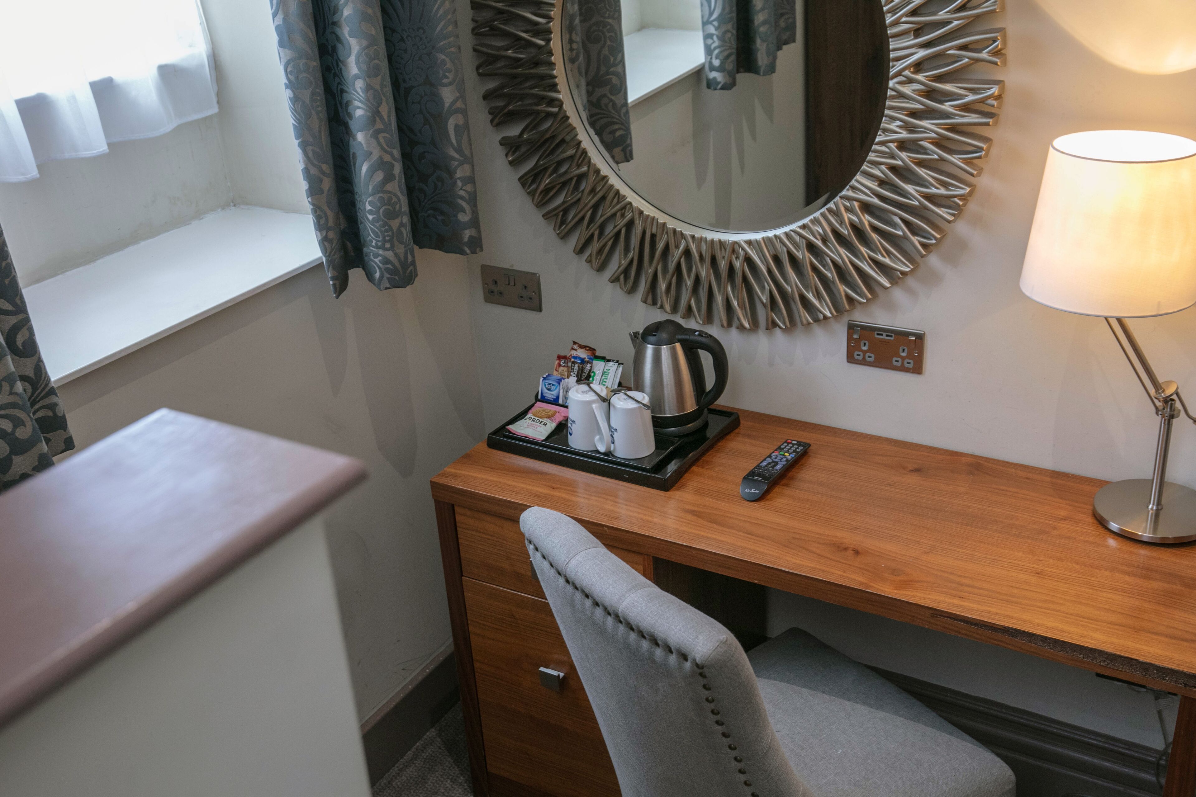 Une chambre d’hôtel avec un bureau en bois, une chaise rembourrée de couleur grise, un miroir rond orné d’un motif en forme de soleil, une lampe de chevet et un plateau contenant des articles de toilette.