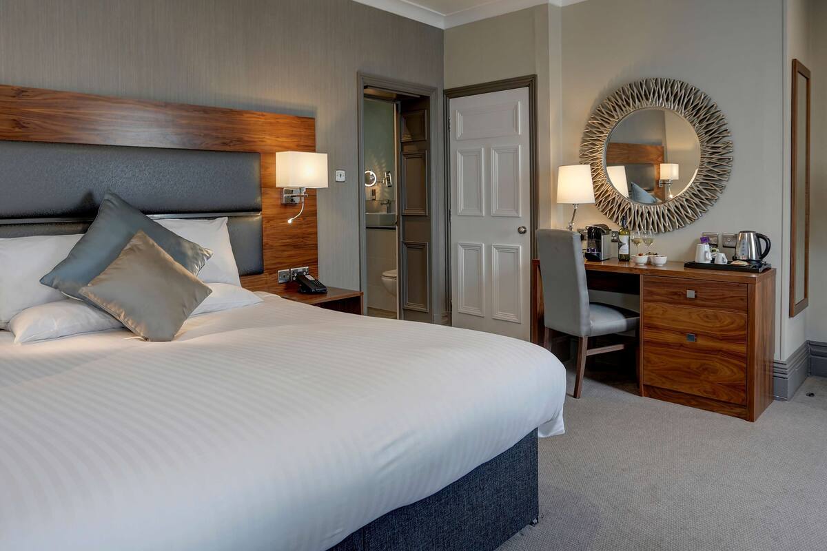 Une chambre d’hôtel avec un grand lit, des lampes de chevet, un bureau avec une chaise, une commode et un grand miroir rond accroché au mur.